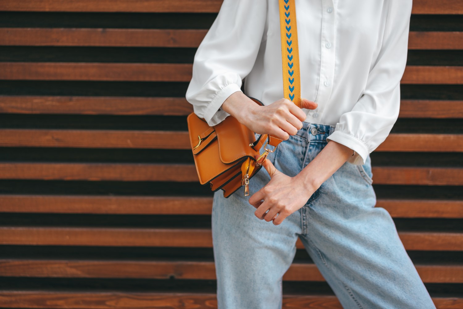 Fashion photo of woman wearing white shirt, jeans and brown crossbody bag against wooden background, close up